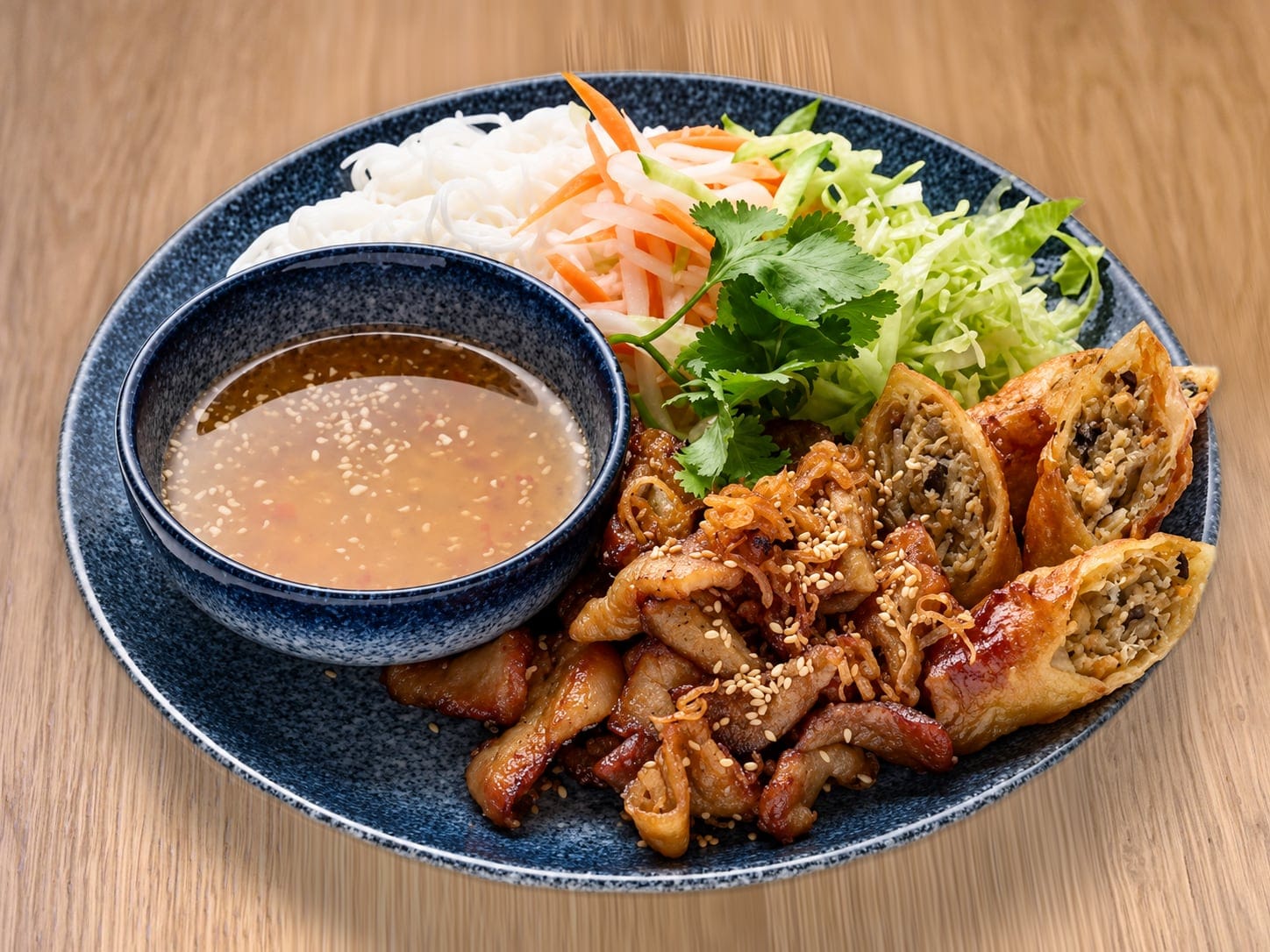 Bún mix plate — rice vermicelli with grilled pork, crispy spring rolls, pickled carrot, lettuce, herbs and a bowl of fish sauce, on a wooden table