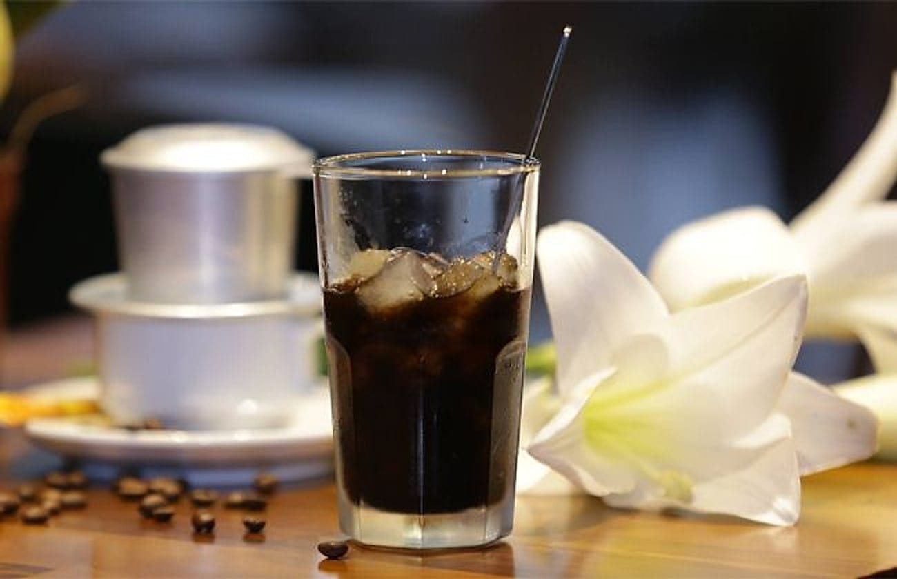 Cà phê đá — iced black Vietnamese coffee beside a phin filter, coffee beans and white lilies on a wooden table