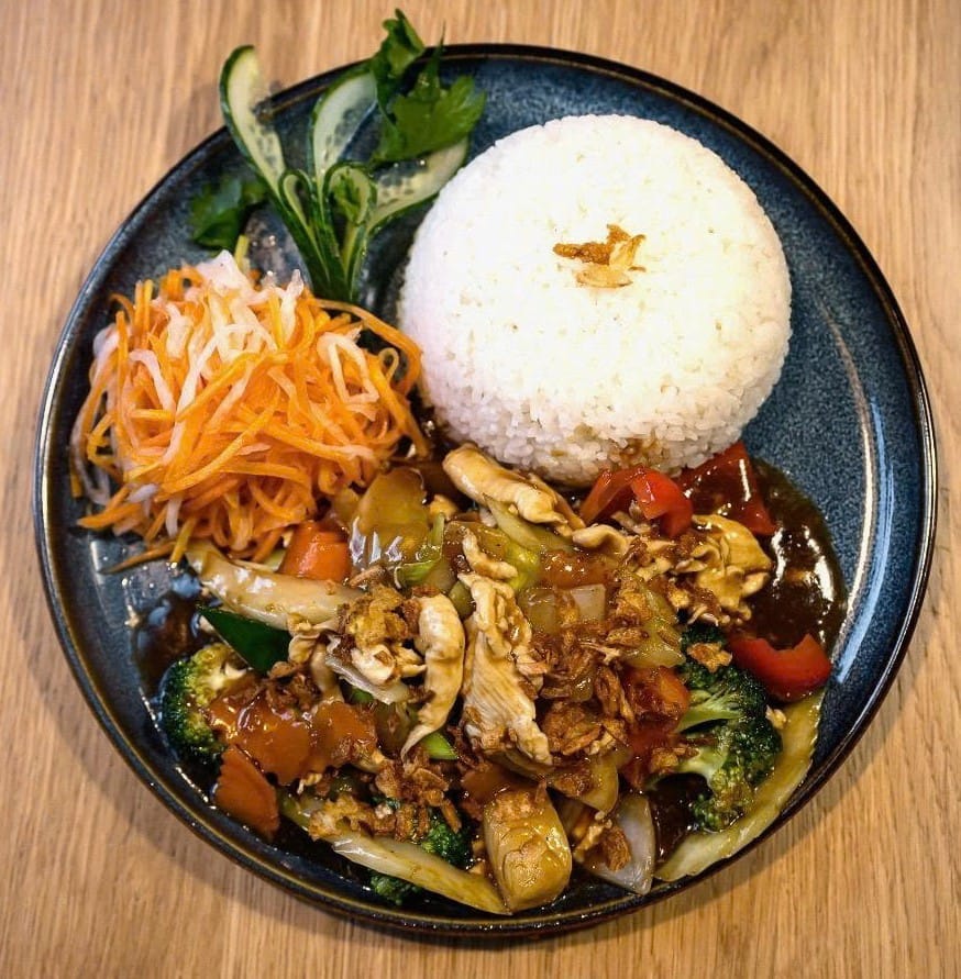 Cơm gà xào — stir-fried chicken with broccoli, peppers, courgette and mushrooms in a savoury sauce, topped with crispy shallots and served with steamed rice, pickled carrot and daikon and cucumber