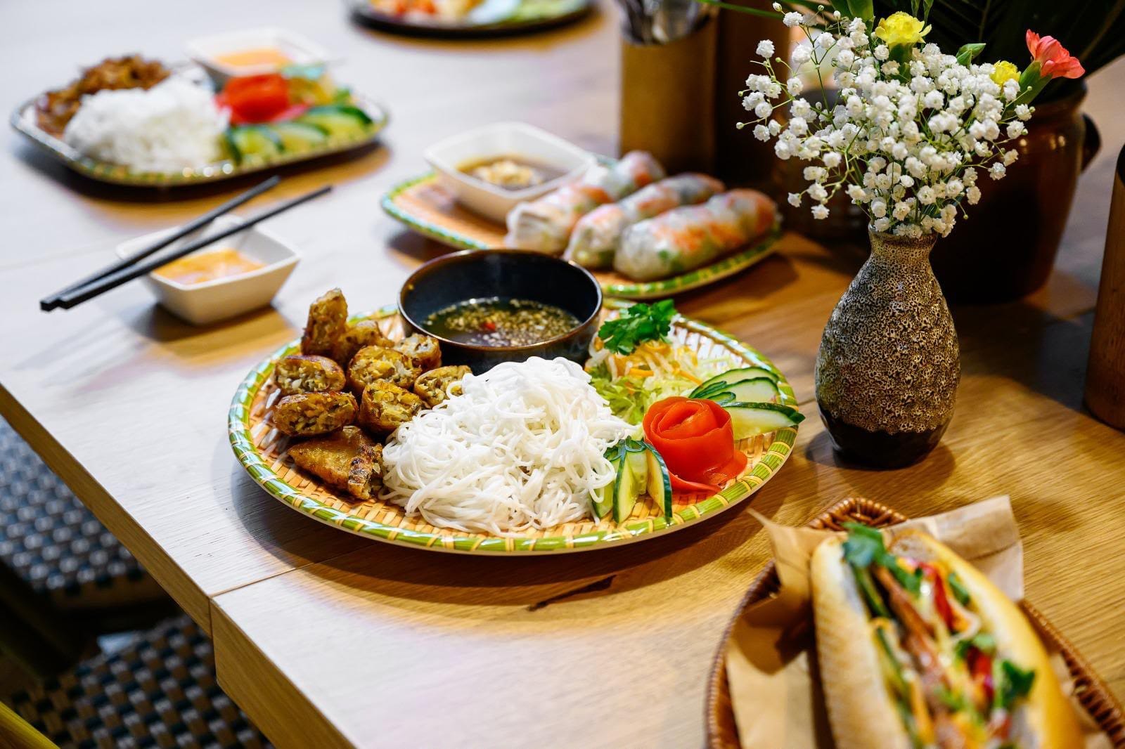 A shared table at Hạ Long Bếp — bún chả nem with crispy spring rolls, gỏi cuốn, bánh mì and a rice plate beside a vase of flowers